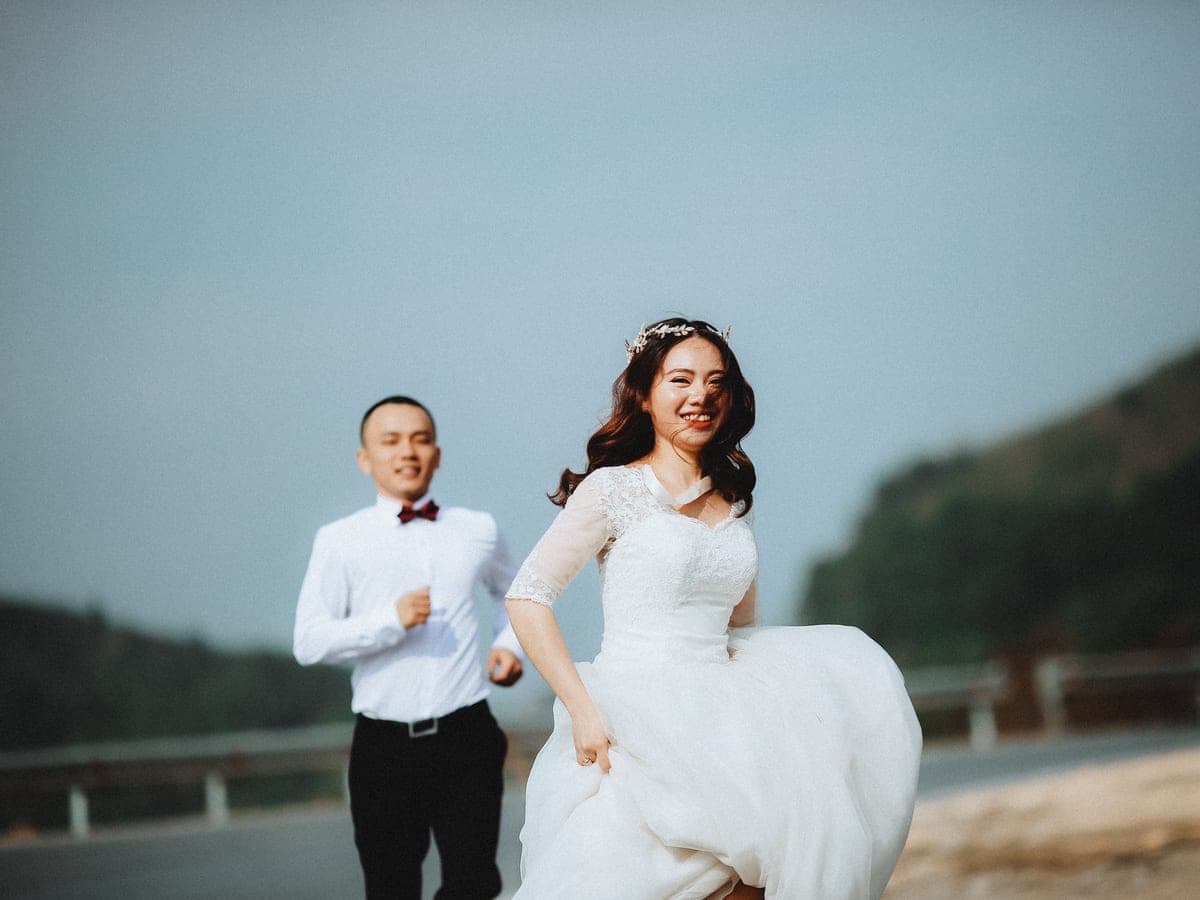 A laughing bride running while the groom follows behind her outdoors.